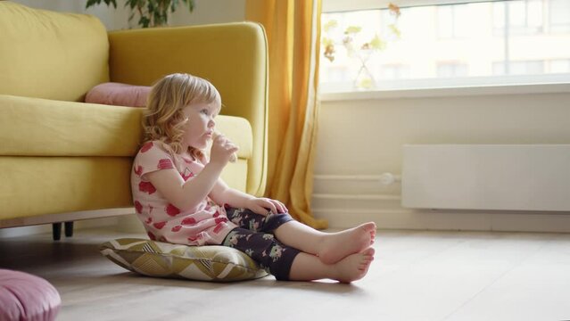 little cheerful girl child in pink dress sits on floor in living room next to yellow sofa, eats banana and watches cartoons on TV. Cute blonde kid enjoys watching TV and eating.