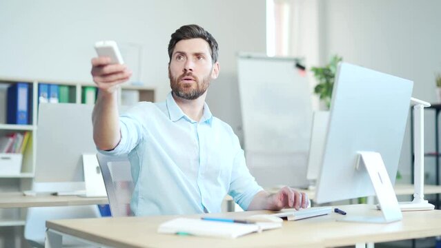 Man employee entrepreneur. A male office worker Turn on a remote control air conditioner, cooling system or humidifier. businessman Sitting at work at a computer desk. Workplace, changing temperature
