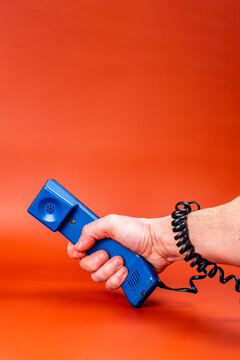 Hand Holding A Blue Phone, With The Phone Cord Wrapped Around The Arm, On An Orange Background.