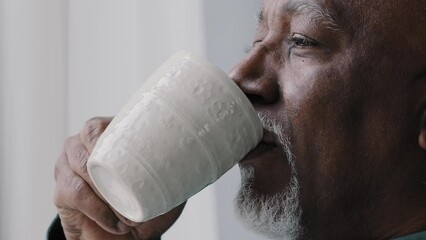 Side view african american old 60s lonely business man drinking coffee tea from cup mug looking out window thinking contemplating daydreaming relaxing enjoying fragrant drink in morning home office