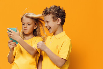 cute, funny children, brother and sister of school age are standing in bright clothes on a yellow background and the girl is holding a phone in a blue case in her hands