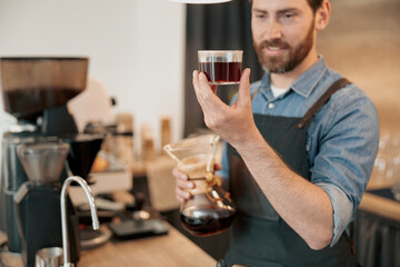 Close up of smiling barista wearing apron appraisingly looks at a glass of filter coffee