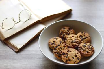 Plate of chocolate chip cookies, open books and reading glasses on wooden table. Selective focus.
