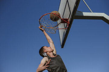 From below of fit sportsman jumping and throwing ball in basketball hoop with rim and net while...