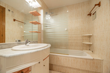Apartment toilet with bathtub with glass partition, cabinet with embedded mirror and lamps, wooden shelves and white and gray marble countertop