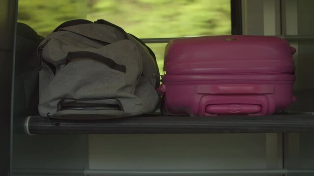 A grey travel bag and a pink suitcase laying on a luggage rack of a train with moving landscape