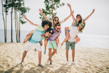 Full length photo of crazy positive friends piggyback raise hands up enjoy warm weather sand beach...