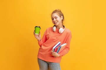 Portrait of young smiling girl, student in casual cloth with books and coffee posing isolated over yellow background
