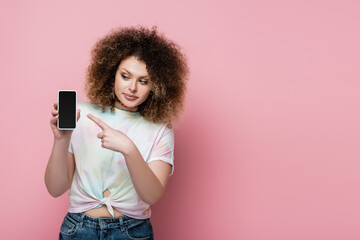 Pretty and curly woman pointing at cellphone isolated on pink.