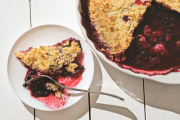 piece of traditional british berry crumble pie, cranberry and raspberry cake on a plate and in baking dish. lifestyle shot.