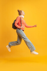 Fototapeta na wymiar Full-length portrait of young girl, student in casual cloth with backpack jumping isolated over yellow studio background