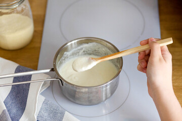 woman's hand holds a wooden spoon of porridge over a pot. Cooking semolina on a white electric stove