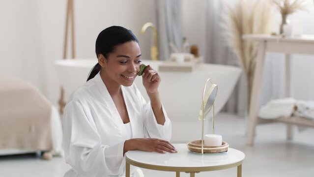 Anti edema morning procedures. Young smiling black lady wearing bathrobe massaging her face with gouache stone
