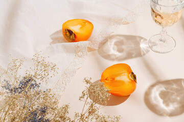 Halfs of persimmon fruits and glasses of water on pastel background with white cloth and sunlit. Summer drinks and refreshment concept.