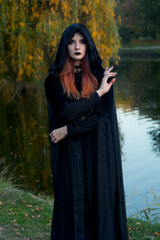 beautiful fashionable young girl witch model stands outdoors in the forest in autumn by the river, pale, vampire, goth