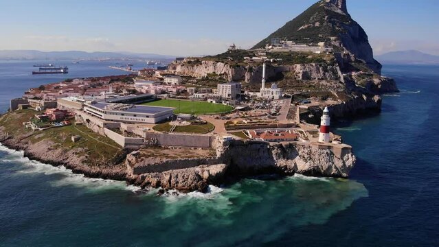 Waves crash against the rocks below the red white colored lighthouse at Europa Point in Gibraltar on a sunny day with in the background the upper rock with great view. Backwards drone dolley shot