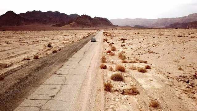 Passenger car drives left-handed fast away on the torn dry dirt road between the red canyon in the dry Timna Park in the Negev Desert in southern Israel on a sunny day. Drone following shot