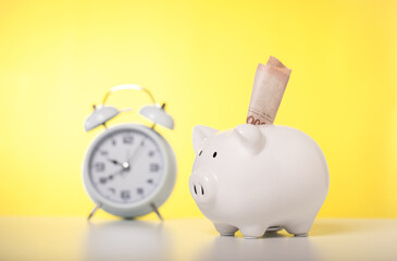 Banknotes are wearing white piggy banks and alarm clock on a table with yellow background, business, investment, finance, and Money Saving for the future concepts, time and money savings.