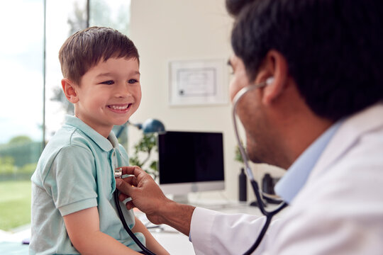 Male Doctor Or GP Wearing White Coat Examining Smiling Boy Listening To Chest With Stethoscope