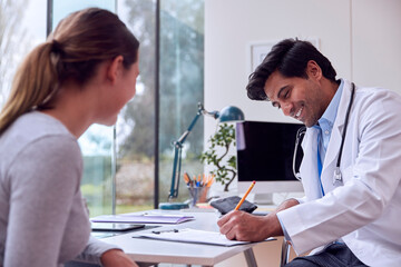 Male Doctor Or GP In Appointment With Teenage Girl Making Notes On Clipboard