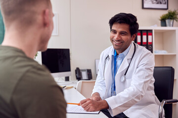 Male Doctor Or GP Wearing White Coat Meeting Young Man For Appointment In Office