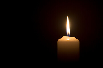 A single burning candle flame or light glowing on a white candle on black or dark background on table in church for Christmas, funeral or memorial service