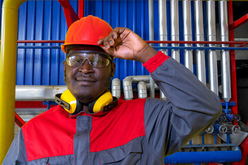 Portrait Of African American Worker In Protective Workwear In Industrial Interior. Industrial Worker In Red Helmet, Protective Eyewear, Hearing Protection Equipment And Work Uniform Looking At Camera.