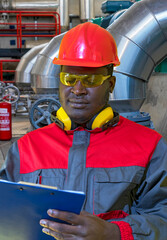 Black Worker In Protective Workwear Writing On Clipboard In Industrial Interior. Young Man In Red Helmet, Protective Eyewear, Hearing Protection Equipment And Work Uniform.