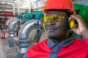 Portrait Of African American Worker In Protective Workwear In Industrial Interior With Pipes And Generators. Worker In Red Helmet, Protective Eyewear, Hearing Protection Equipment And Work Uniform.
