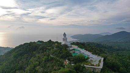 Fototapeta na wymiar Drone view of the Big Buddha, Thailand. The Big Buddha is sitting on hill in the lotus position, meditating. The sun shines brightly through clouds. There's a jungle all around. A ray of sun on water