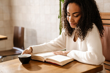 Young black woman drinking coffee and reading book in cafe