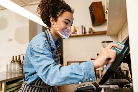 Black barista woman wearing face mask working with cash register in cafe