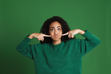 Young black woman grimacing while pointing fingers at her cheeks