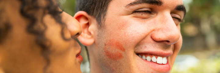 Multiethnic Young people kiss on cheek with lipstick – portrait of two people kissing on cheek – woman kissing her boyfriend -  happy loving woman kissing boy friend on cheek   - obrazy, fototapety, plakaty