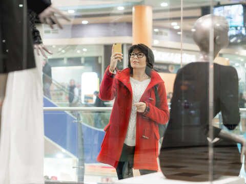 Woman takes photos of dummies in clothing store. Casual outfit hanging on mannequins. View through transparent shop window. Shopping at mall. Basic clothes for everyday wear. Modern urban fashion.
