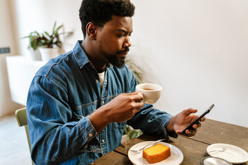 Fototapeta na wymiar Black bearded man using mobile phone while drinking tea in cafe