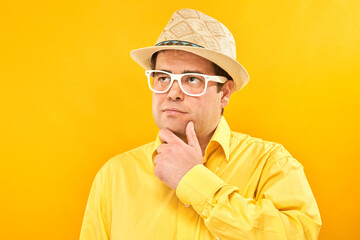Positive man in panama hat holding chin thinks about vacation, doubts, makes decision isolated on yellow studio background