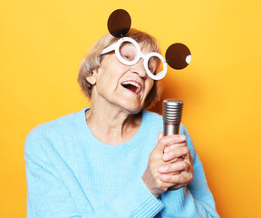 Happy old woman singing with microphone over yellow color background, having fun