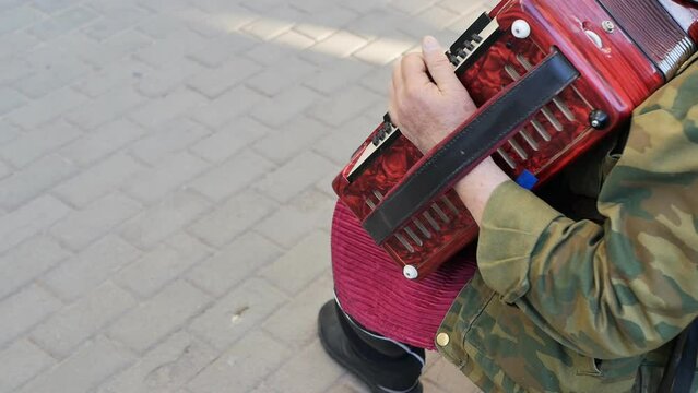 Caucasoid senior man street musician plays his music on the harmonica in the city square in the open air. The hand of a street elderly musician presses the keys of the accordion and stretches the furs