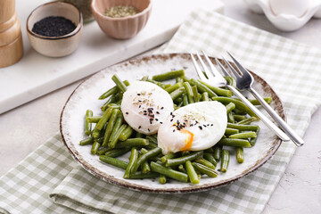 Soft-boiled poached eggs with black sesame seeds on a cooked green beans on a plate with brown rim on a green checkered kitchen towel