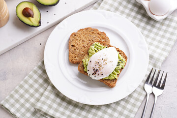 Slices of gluten-free sunflower seeds bread with mashed avocado, poached egg and sesame seeds on white plate on green checkered napkin