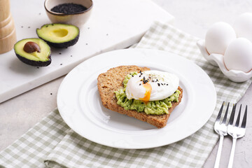 Slices of gluten-free sunflower seeds bread with mashed avocado, poached egg and sesame seeds on white plate on green checkered napkin