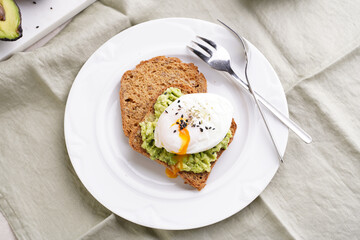 Slices of gluten-free sunflower seeds bread with mashed avocado, poached egg with egg yolk dripping and sesame seeds on white plate on green checkered napkin
