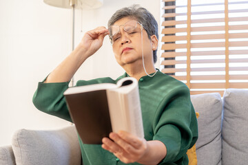 Presbyopia, Hyperopia mature asian woman holding eyeglasses having problem with vision problem trying to read text on book, eye disease of old elderly sitting on couch. Poor eyesight, health care.