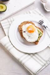 Slices of gluten-free sunflower seeds bread with mashed avocado, fried egg and sesame seeds on white plate on green checkered napkin