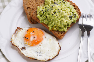 Slices of gluten-free sunflower seeds bread with mashed avocado, fried egg and sesame seeds on white plate on green checkered napkin