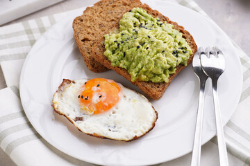 Slices of gluten-free sunflower seeds bread with mashed avocado, fried egg and sesame seeds on white plate on green checkered napkin