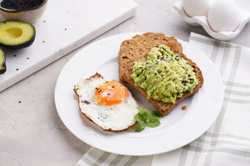 Slices of gluten-free sunflower seeds bread with mashed avocado, fried egg and sesame seeds on white plate on green checkered napkin