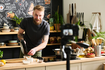 Young Caucasian man with beard on face making content for food blog pouring fried sesame seeds into bowl - obrazy, fototapety, plakaty