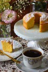 Fatia de bolo de fubá coberto de açúcar em cenário aconchegante de um café da manhã, com flores ao redor.